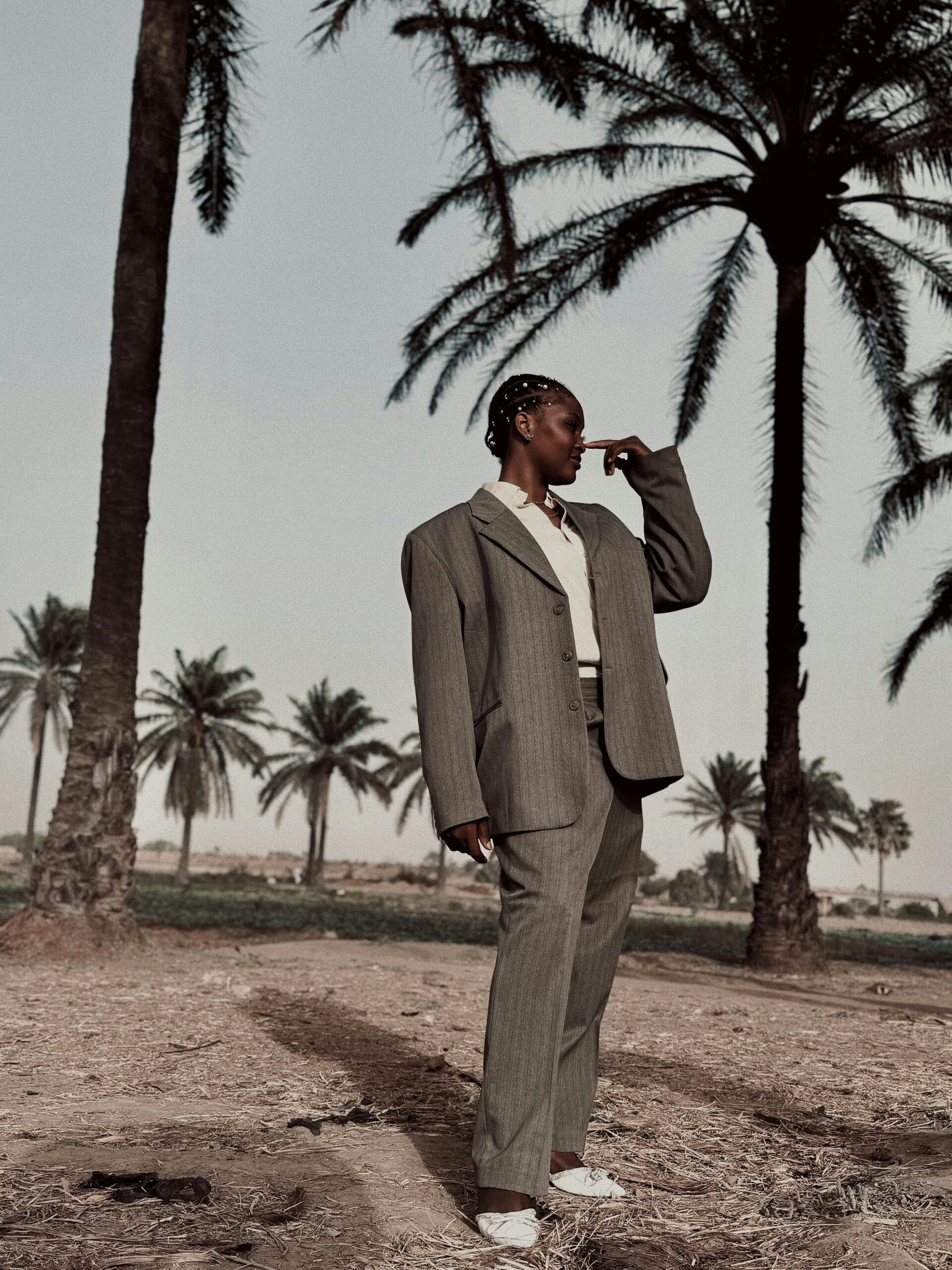 Fashionably dressed woman in a suit poses amidst palm trees in a desert setting.